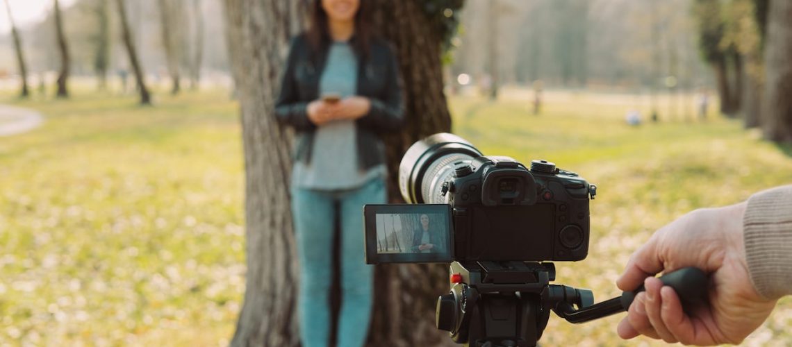 Video shooting at the park: video camera and operator hand on foreground and female model standing in the background